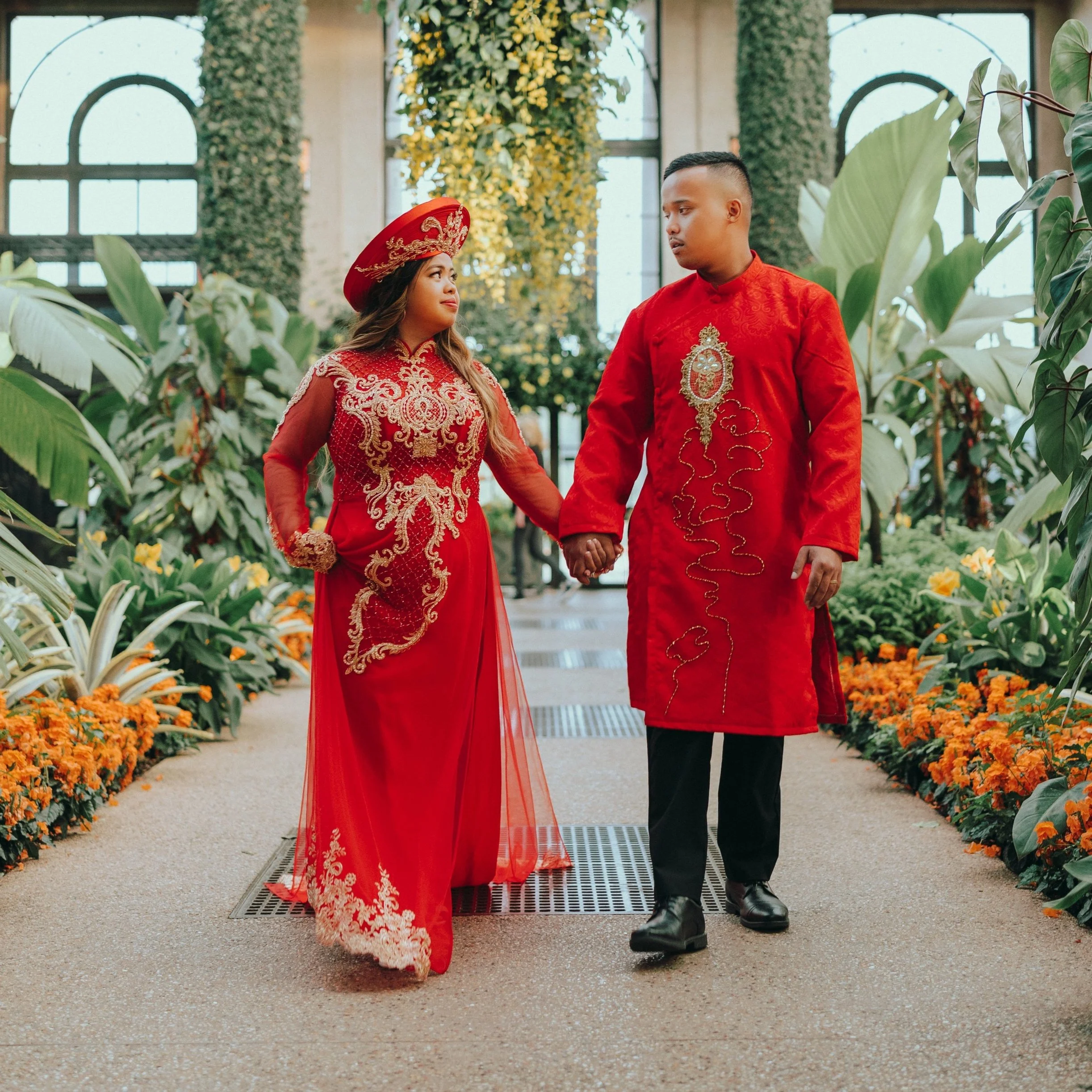 A couple in traditional Vietnamese wedding attire holding hands and walking through a lush indoor garden with vibrant orange flowers and green foliage.