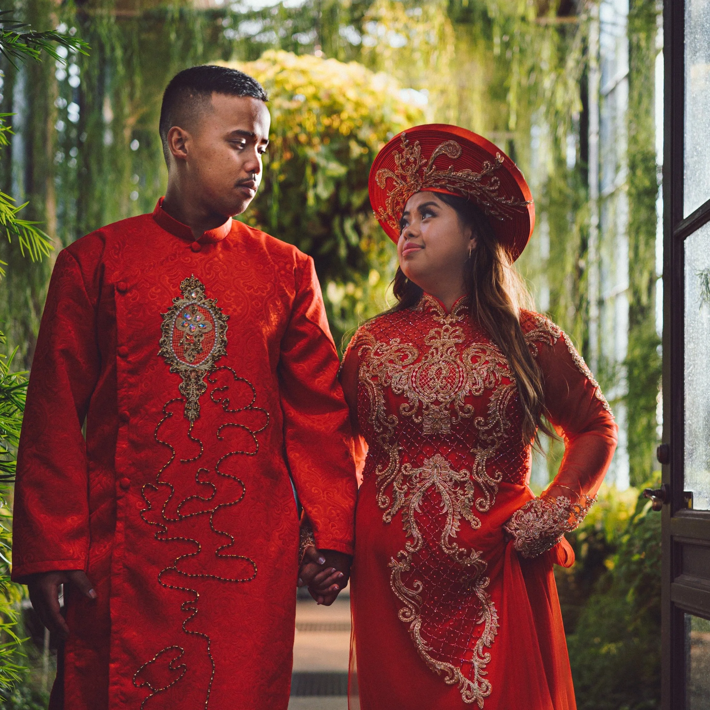 A couple in traditional Indonesian wedding attire holding hands in a lush greenhouse or botanical setting.