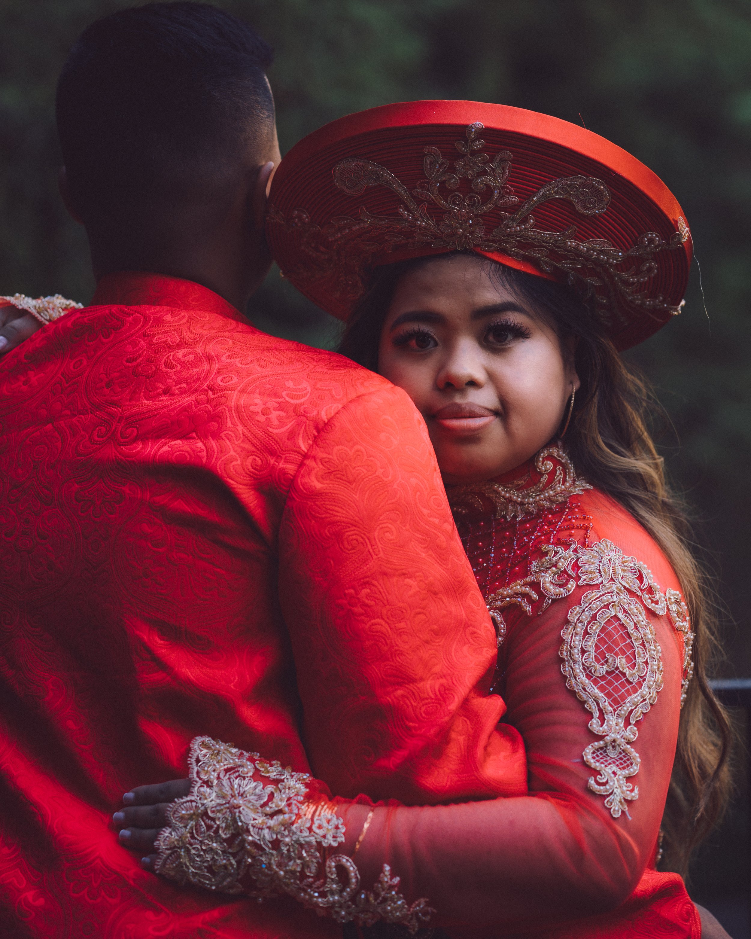 A woman in traditional Mexican attire embraces a man, wearing a red embroidered dress and a large red sombrero, during a cultural celebration or wedding.