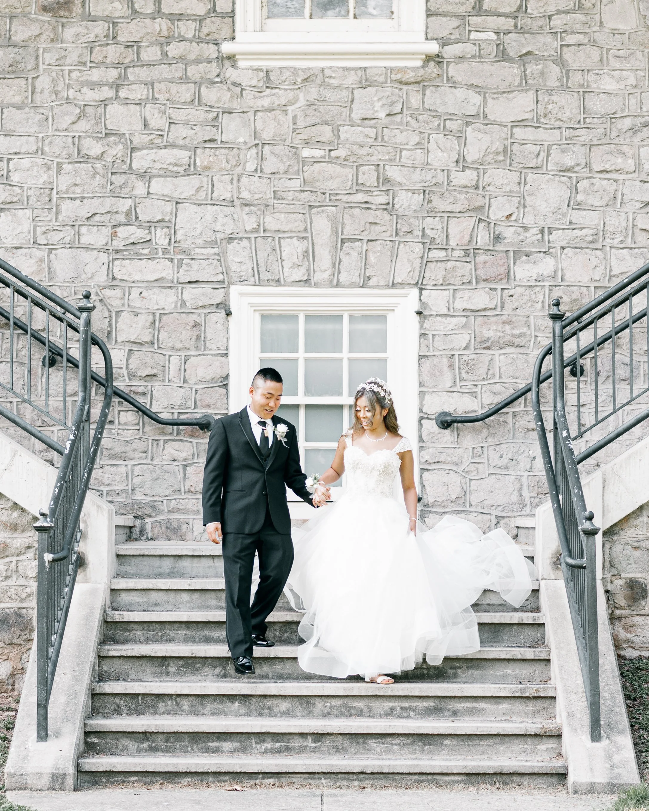 A bride and groom stepping down gray concrete stairs with black metal railings outside a stone building. The bride is wearing a white wedding gown with a floral headpiece, and the groom is dressed in a black suit with a white shirt and black tie.