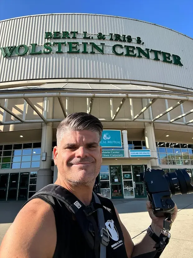 Damien Campbell taking a selfie in front of Wolstein Center at Cleveland State University, with a camera in his hand and a bright blue sky above.