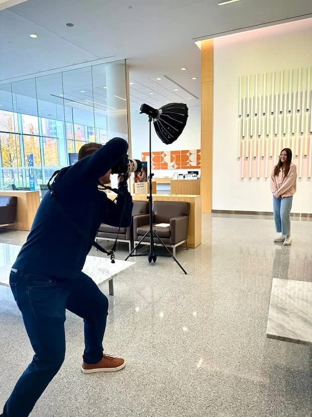 Photographer Damien Campbell is taking pictures of a young woman who is posing in a well-lit lobby area with modern decor, including colorful wall art and seating.
