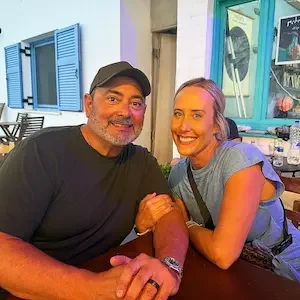A man and a woman sitting together at a table, smiling. The man is wearing a black shirt and cap, and the woman is in a gray top with tattered sleeves. They are outdoors, with colorful buildings and window shutters in the background.