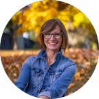 A smiling woman with glasses in a denim jacket outdoors with fall foliage in the background.