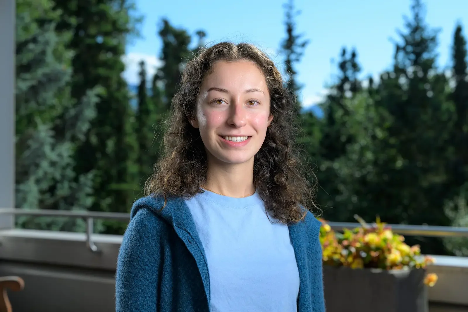 A woman with curly brown hair wearing a light blue shirt and a dark blue cardigan, smiling outdoors on a balcony with trees and blue sky in the background.