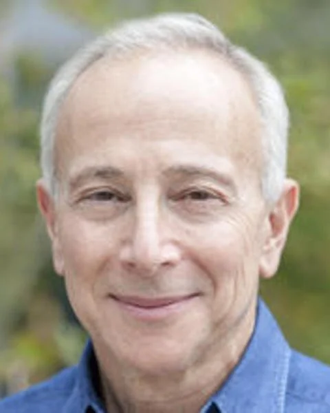 Older person with short light gray hair smiling softly, wearing a blue collared shirt, with greenery blurred in the background.