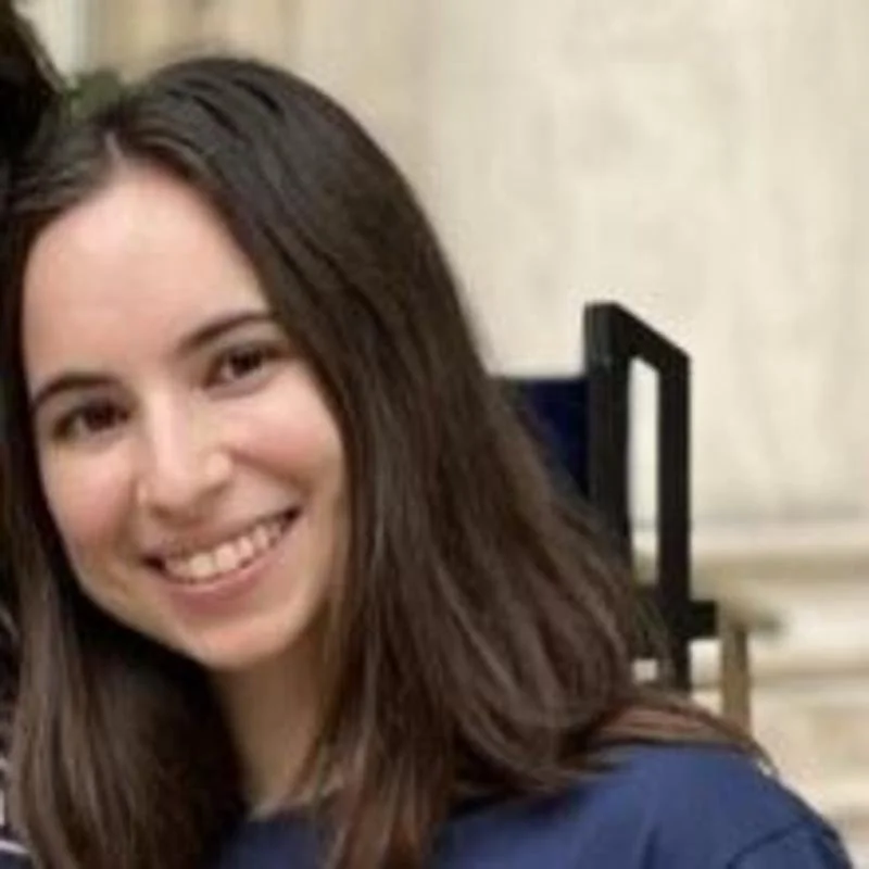 Person with long straight dark hair smiling slightly, wearing a dark top, with an indoor setting softly blurred in the background.