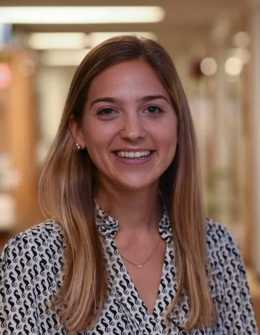 Person with straight, shoulder‑length light brown hair smiling, wearing a patterned blouse and small earrings, standing in a warmly lit hallway.