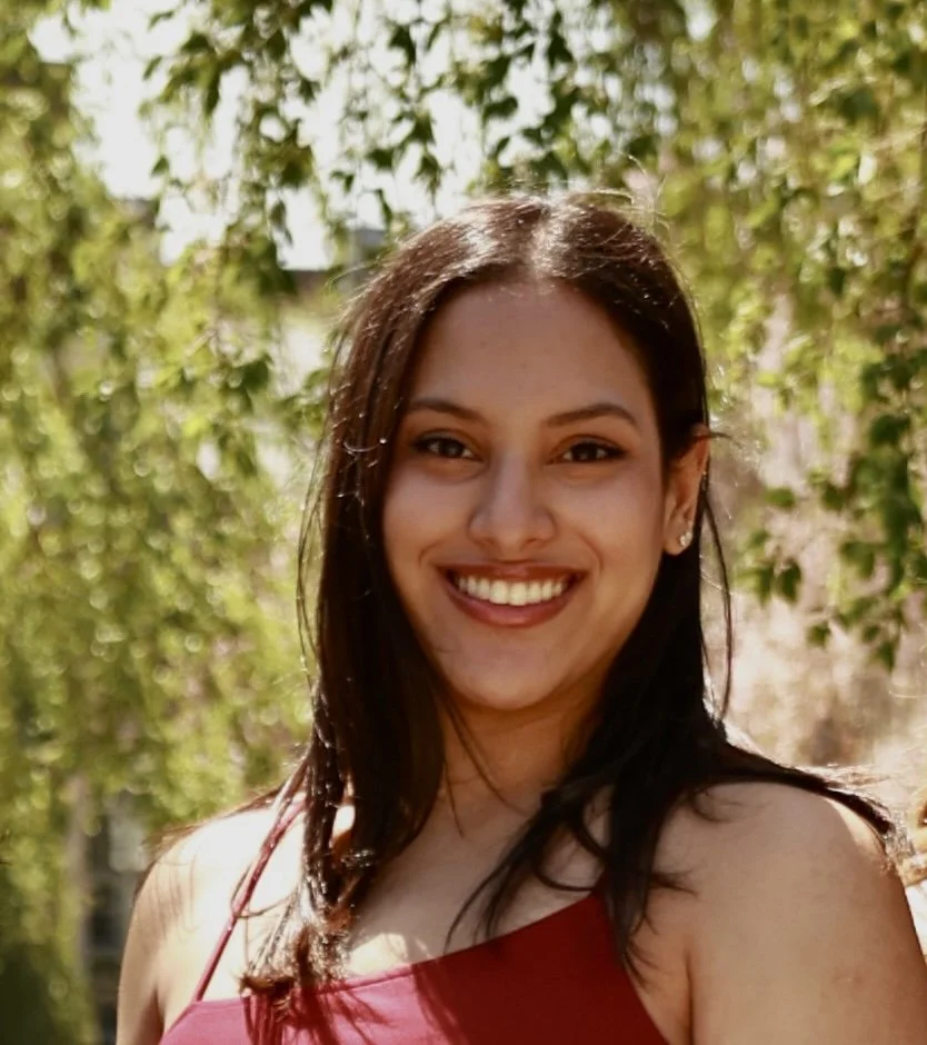 Smiling woman with straight dark hair wearing a red sleeveless top, standing outdoors in front of sunlit green foliage.