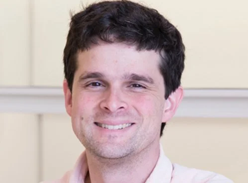 Head-and-shoulders photo of a smiling man with short dark hair, light skin, and a light-colored collared shirt, posed indoors against a softly blurred beige background.