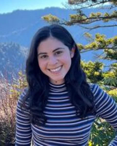 A smiling woman with long, dark hair wearing a navy blue, white, and pink striped long-sleeve shirt, standing outdoors with evergreen trees and blue mountains in the background.