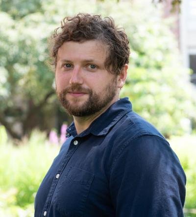 Person with curly light brown hair and beard standing outdoors, wearing a dark blue button‑up shirt, looking at the camera with greenery softly blurred in the background.