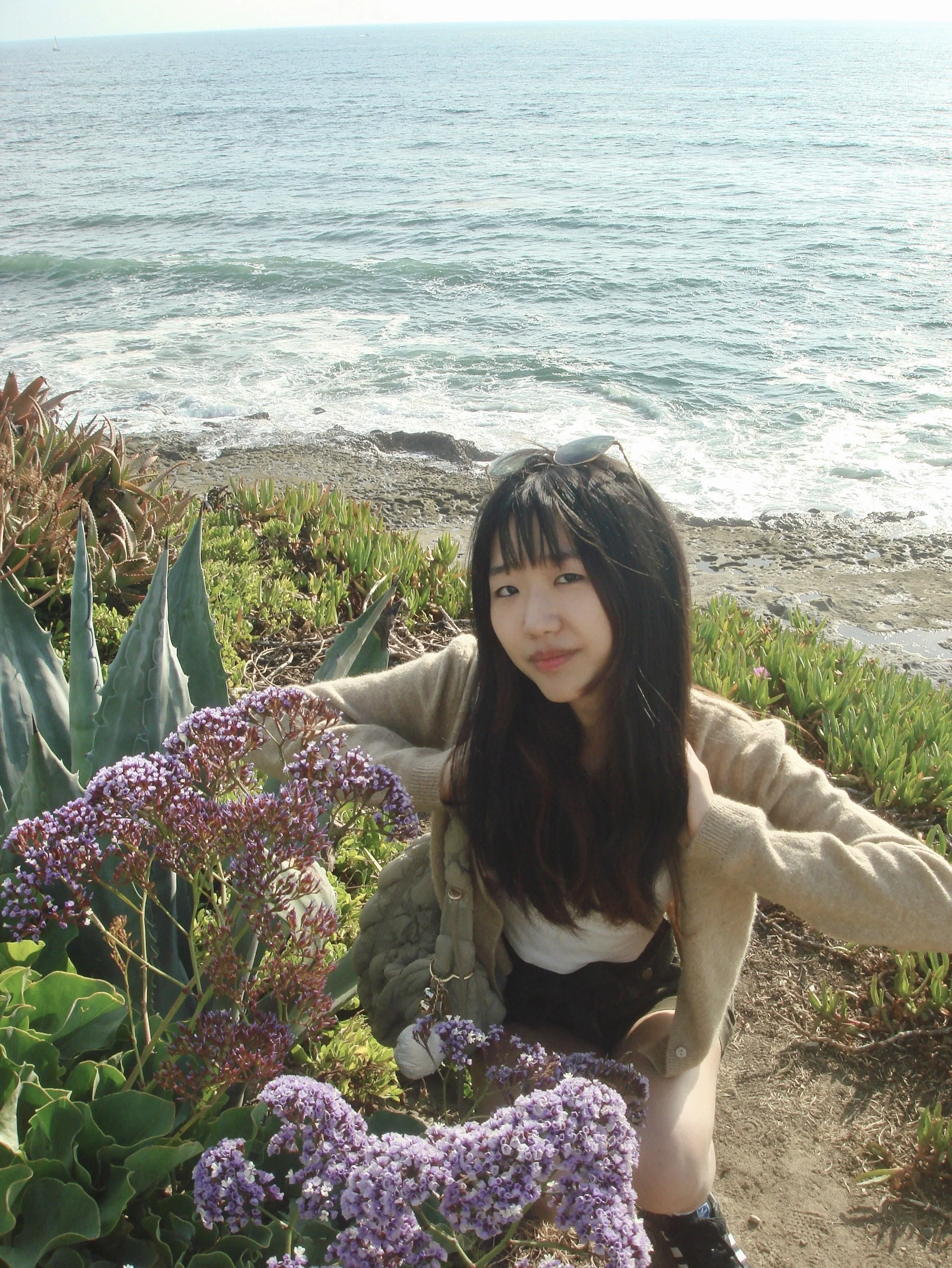 Young woman with long dark hair and bangs crouches beside purple flowering plants on a coastal bluff. She wears a tan cardigan, light top, and shorts, with the ocean and rocky shoreline visible behind her.