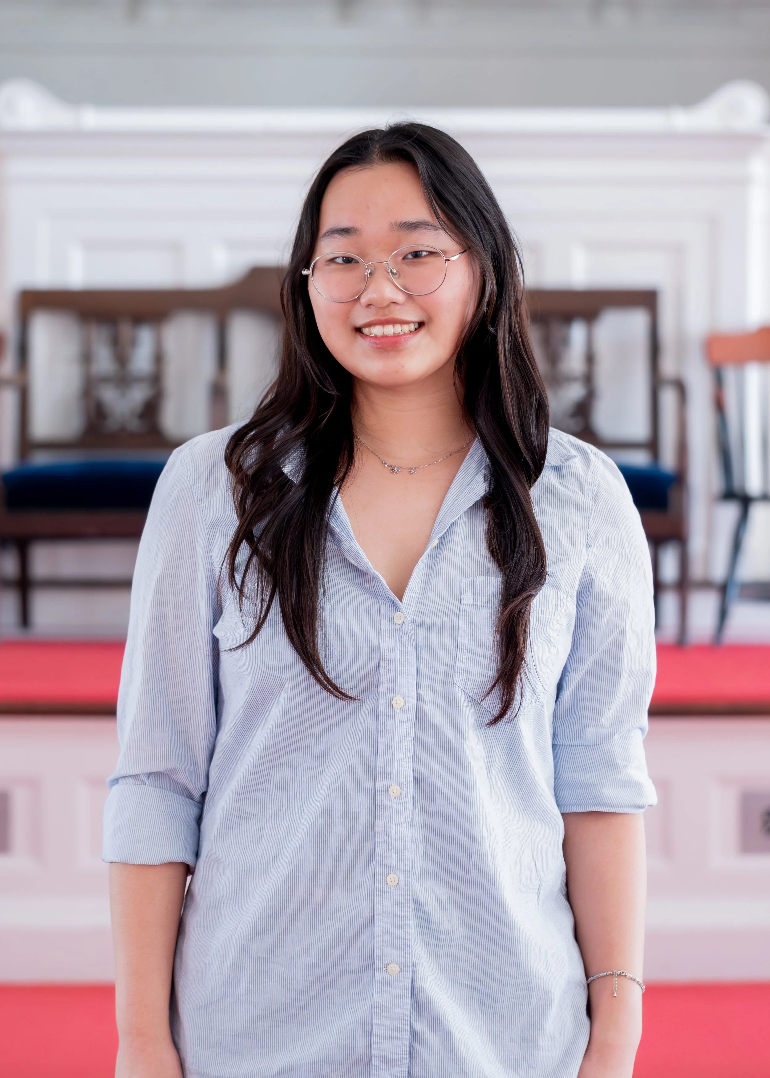 Young woman with long dark hair and glasses stands indoors in front of wooden chairs and a white wall. She wears a light blue button‑down shirt with rolled‑up sleeves and a small necklace, smiling at the camera.