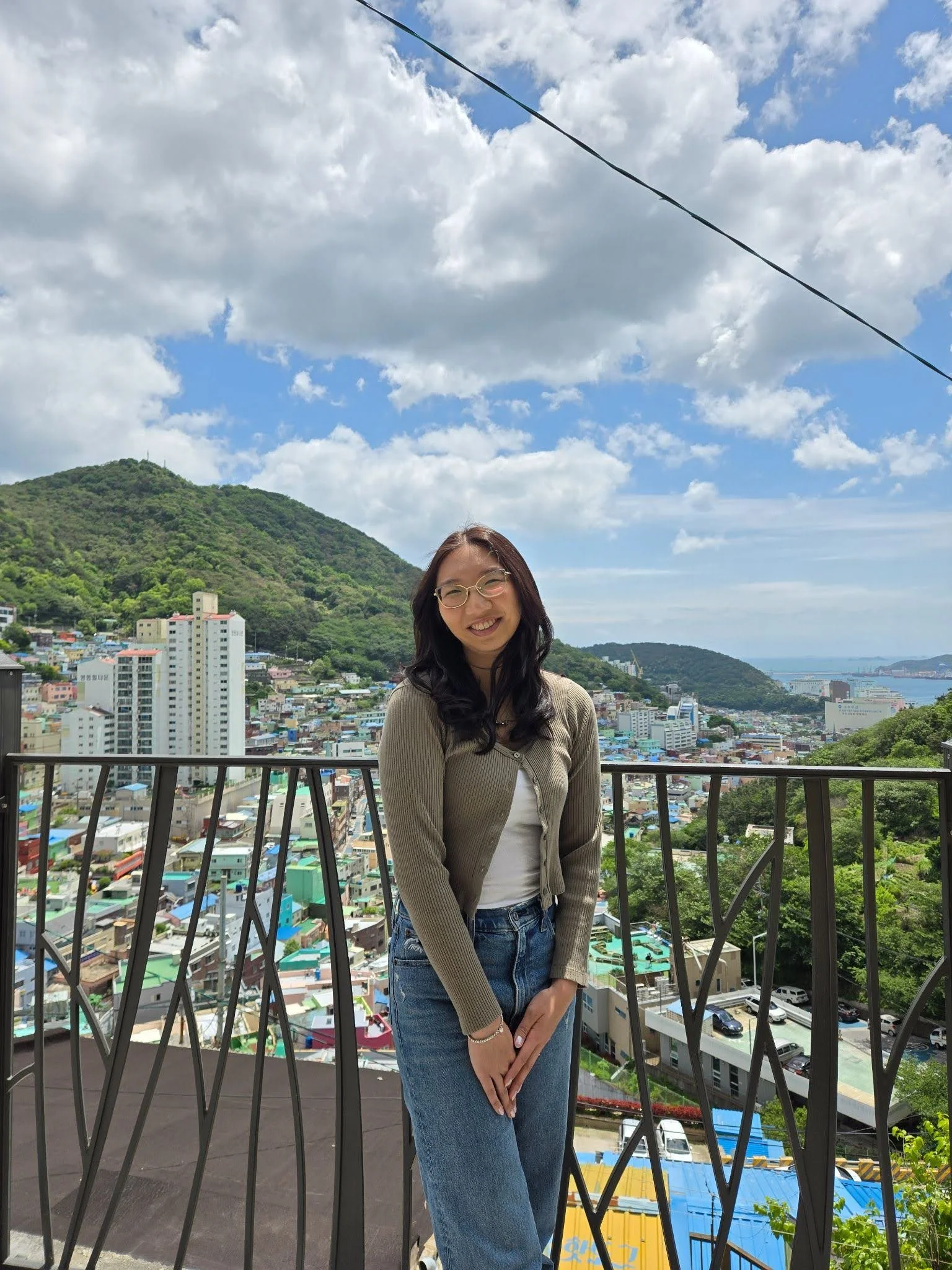 A person with long dark hair and glasses stands smiling on a balcony, wearing a light brown cardigan and jeans. Behind them is a colorful hillside city with green mountains and a bright blue sky filled with fluffy clouds.