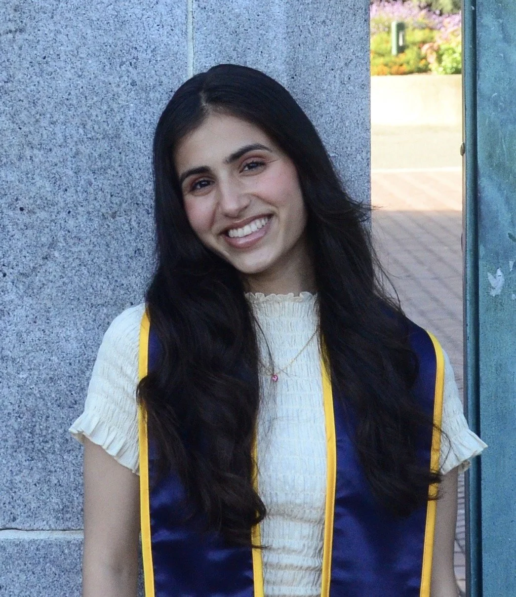 Person with long dark wavy hair smiles at the camera, wearing a light textured dress and a dark graduation stole with yellow trim, standing against a stone wall outdoors.