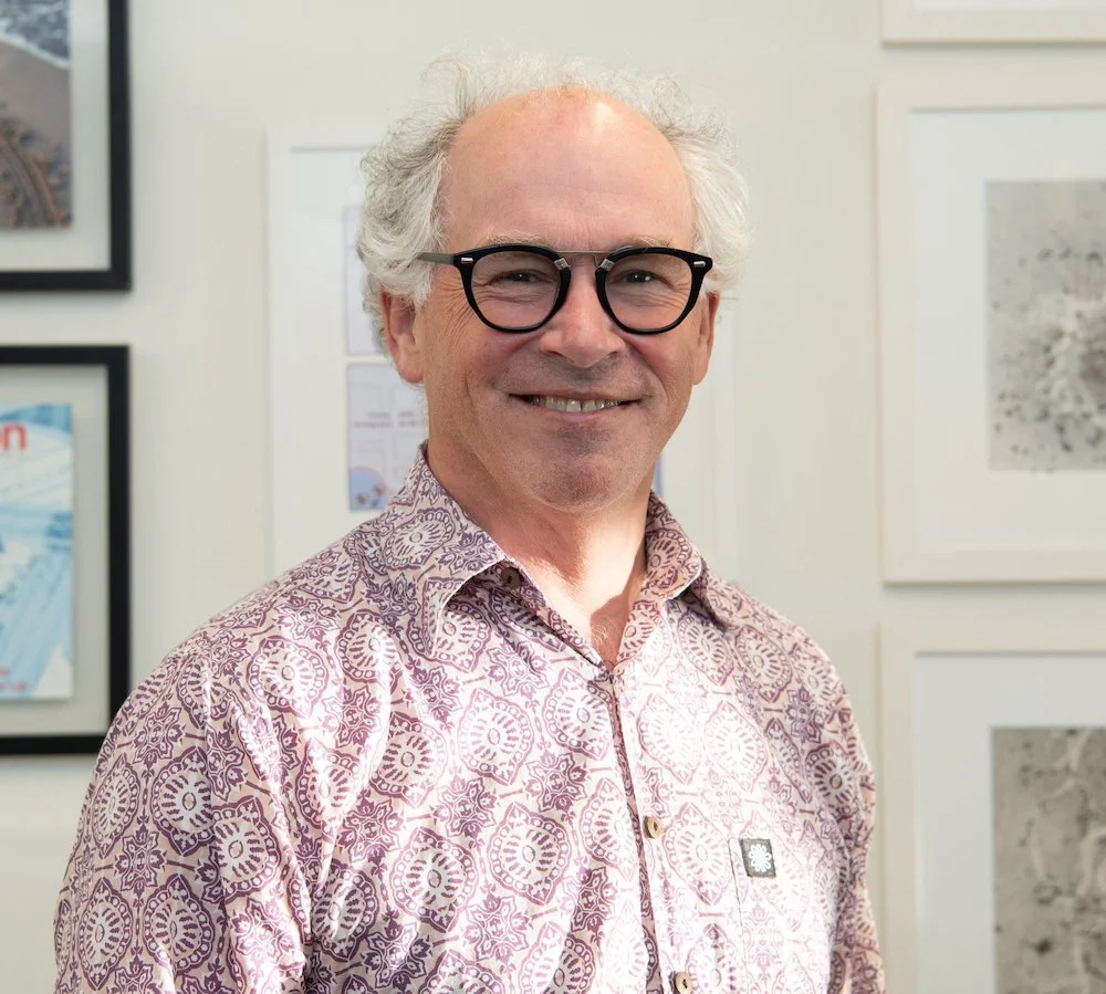 Middle‑aged person with white hair and black glasses smiling, wearing a patterned pink button‑up shirt, standing in a bright room with framed artwork on the walls.