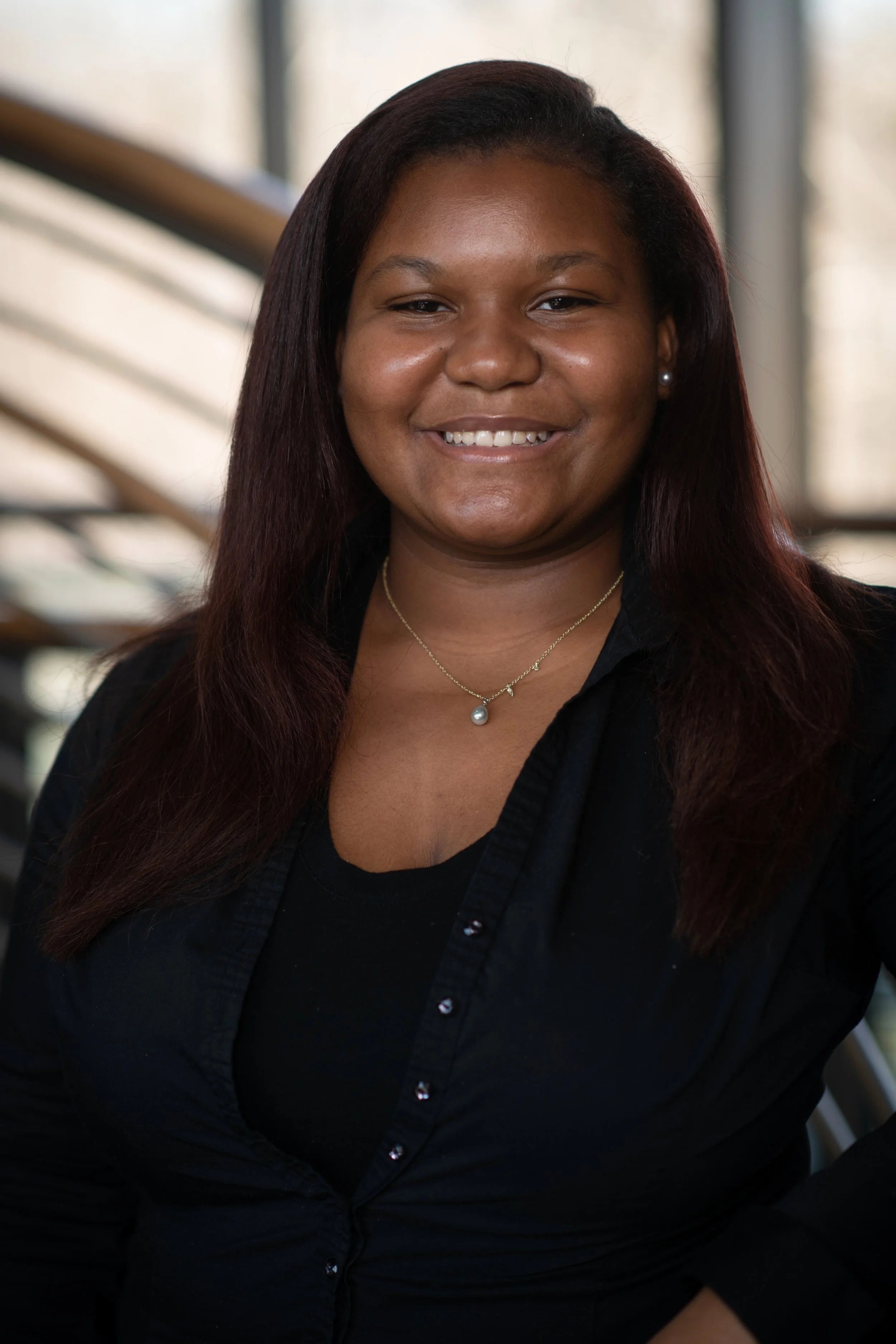 A woman with straight shoulder-length hair, wearing a black top and a pearl necklace, smiling in an indoor setting with windows in the background.