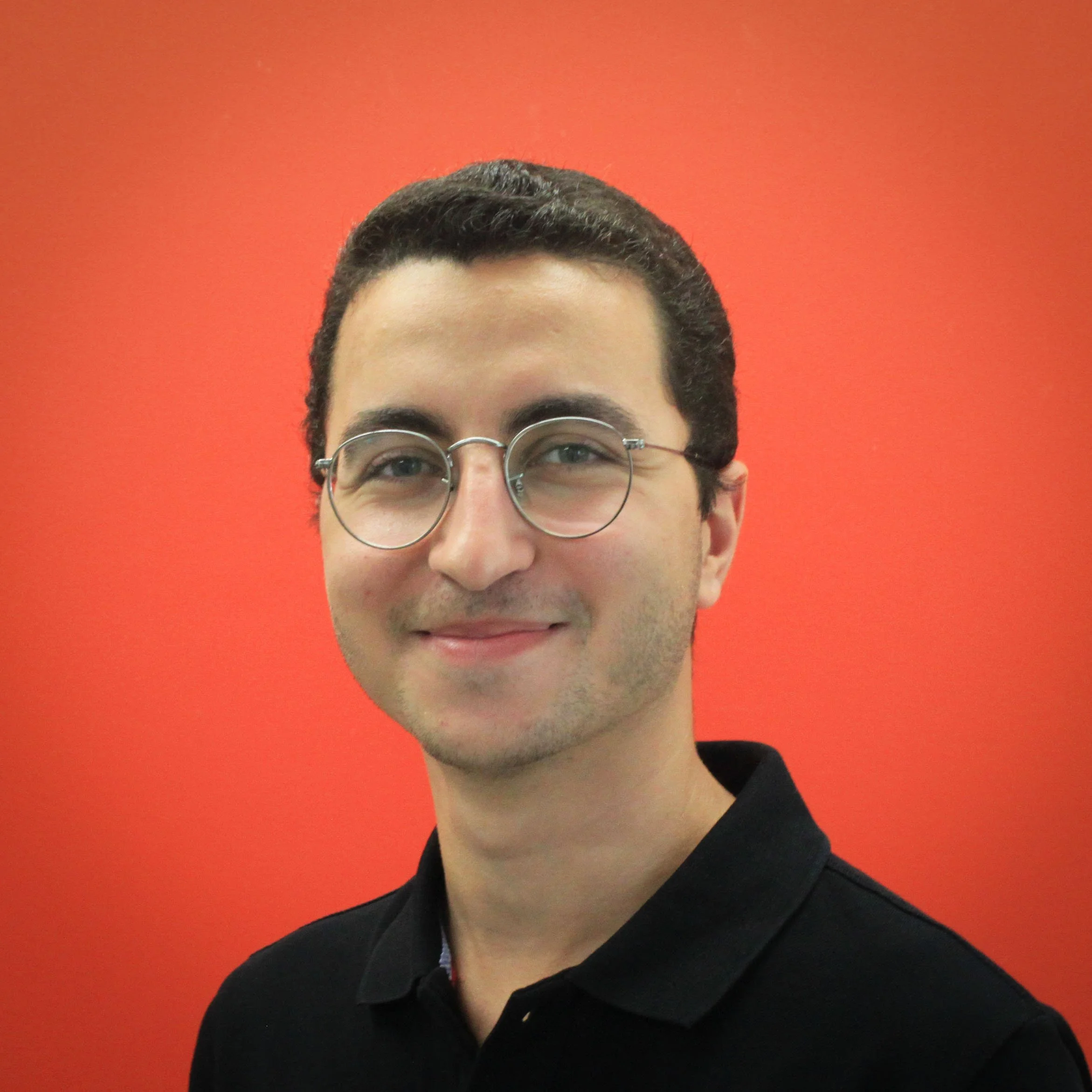 A person with short dark hair and round glasses smiles at the camera in front of a solid bright orange background, wearing a black collared shirt.