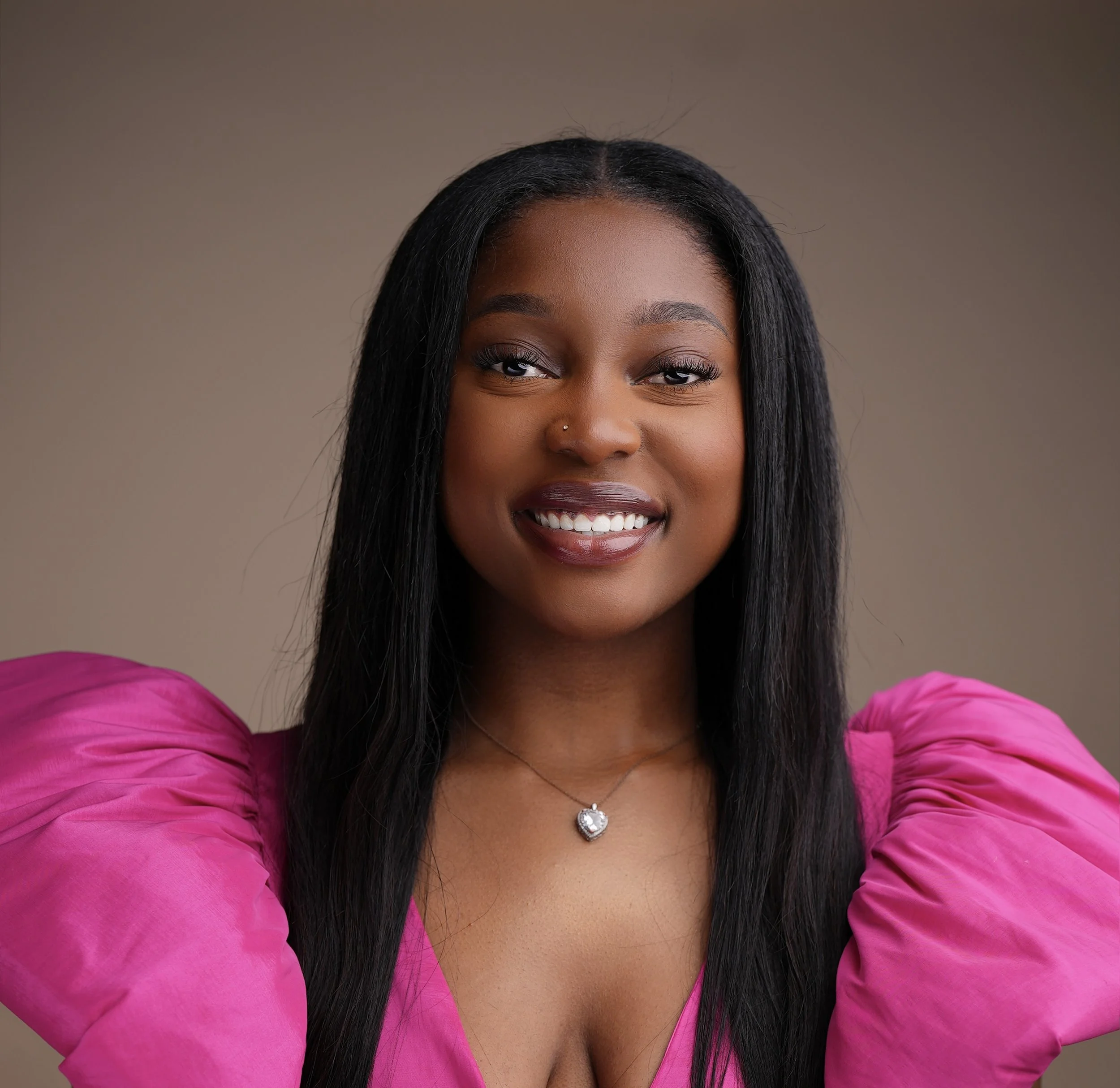 A woman with long straight hair, wearing a bright pink puff-sleeve dress and a heart-shaped pendant necklace, smiling in front of a plain background.