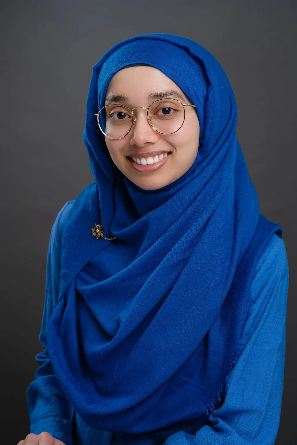A person wearing glasses and a bright blue hijab and top smiles at the camera. The background is a simple dark gray studio backdrop.
