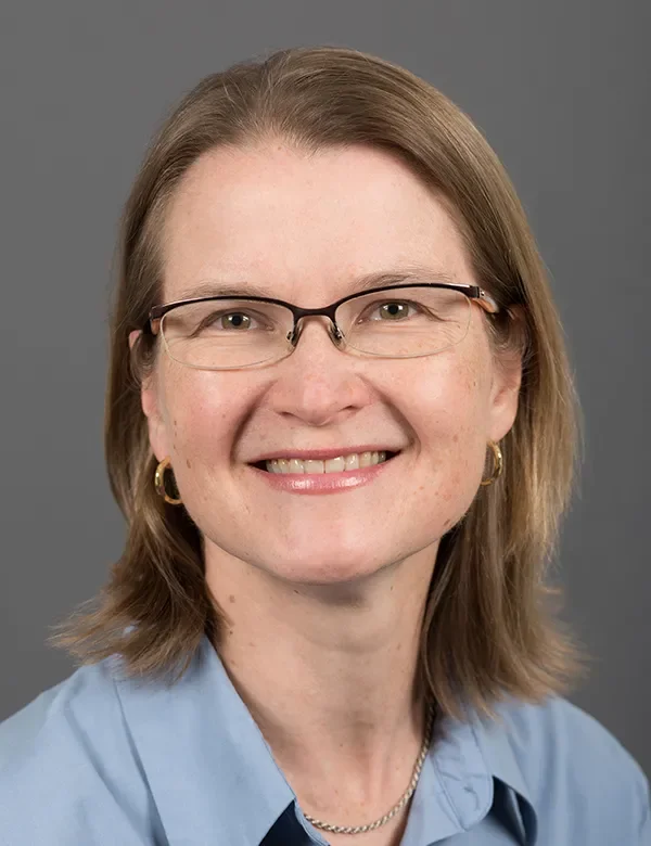 A person with straight, shoulder-length light brown hair and glasses smiles at the camera, wearing small hoop earrings and a light blue collared shirt against a plain gray background.