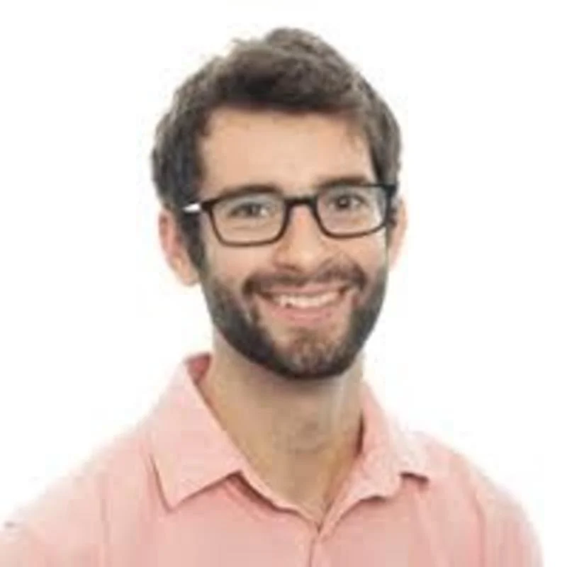 A person with short dark hair, glasses, and a beard smiles at the camera. They are wearing a light pink collared shirt against a plain white background.