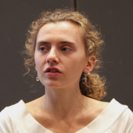 A young person with curly blond hair pulled back is speaking, looking slightly to the side, against a dark indoor background. They are wearing a white top and small dangling earrings.