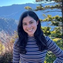 A young adult with long dark wavy hair smiles at the camera, standing outdoors in front of sunlit evergreen trees and distant blue mountains. They wear a long‑sleeve, black-and-white striped turtleneck shirt.