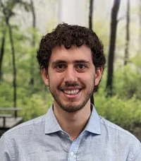 A person with short curly dark hair and a beard smiles at the camera. They are wearing a light blue button-up shirt, with a softly focused background of tall trees and greenery.
