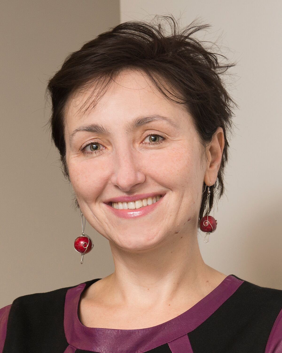 A person with short dark hair and dangling red earrings smiles warmly at the camera, wearing a black top with purple trim, standing in front of a neutral indoor background.
