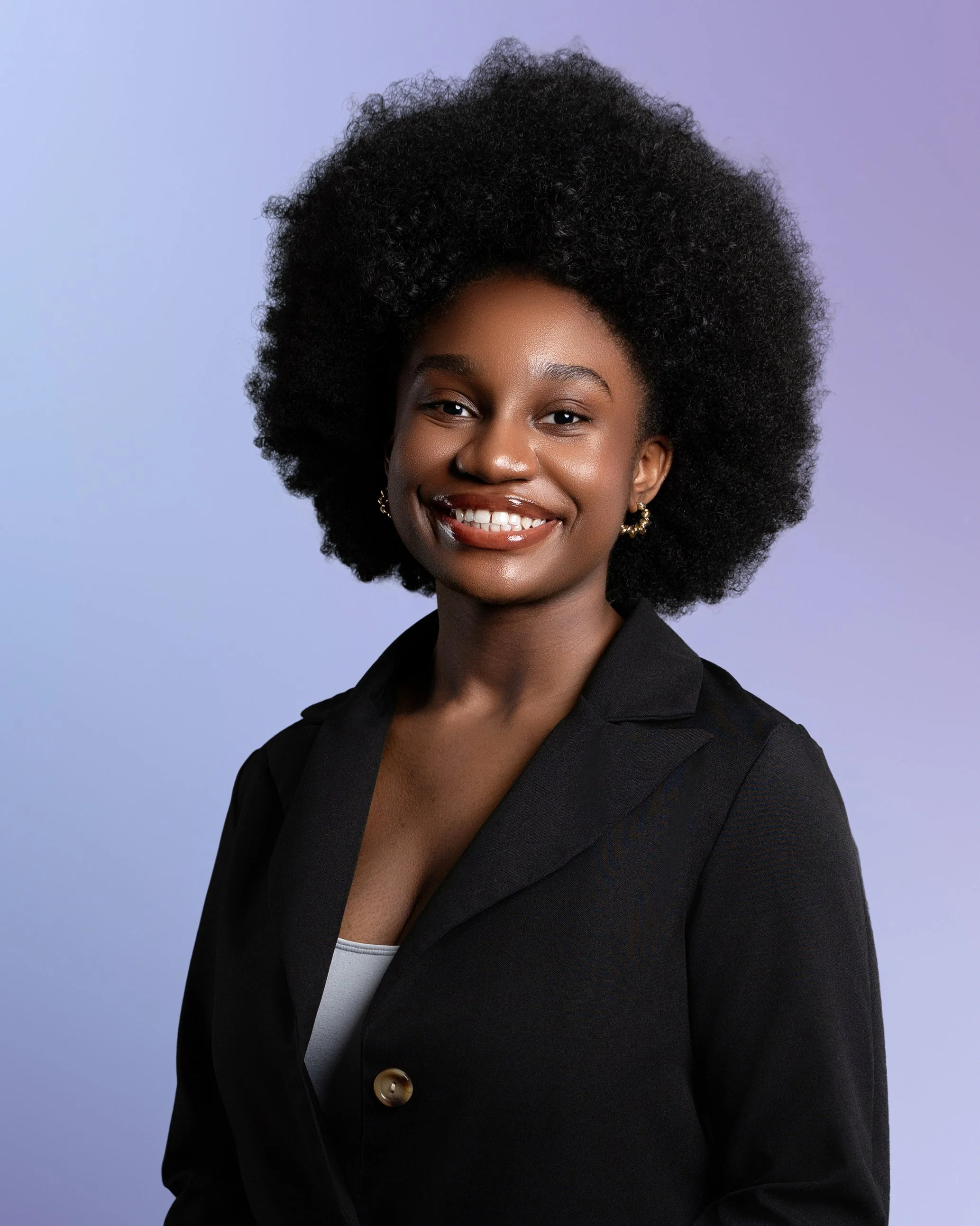 Studio portrait of a young woman with a large natural afro hairstyle, smiling brightly. She wears gold hoop earrings, a black blazer with a single gold button, and a light top, set against a soft purple gradient background.