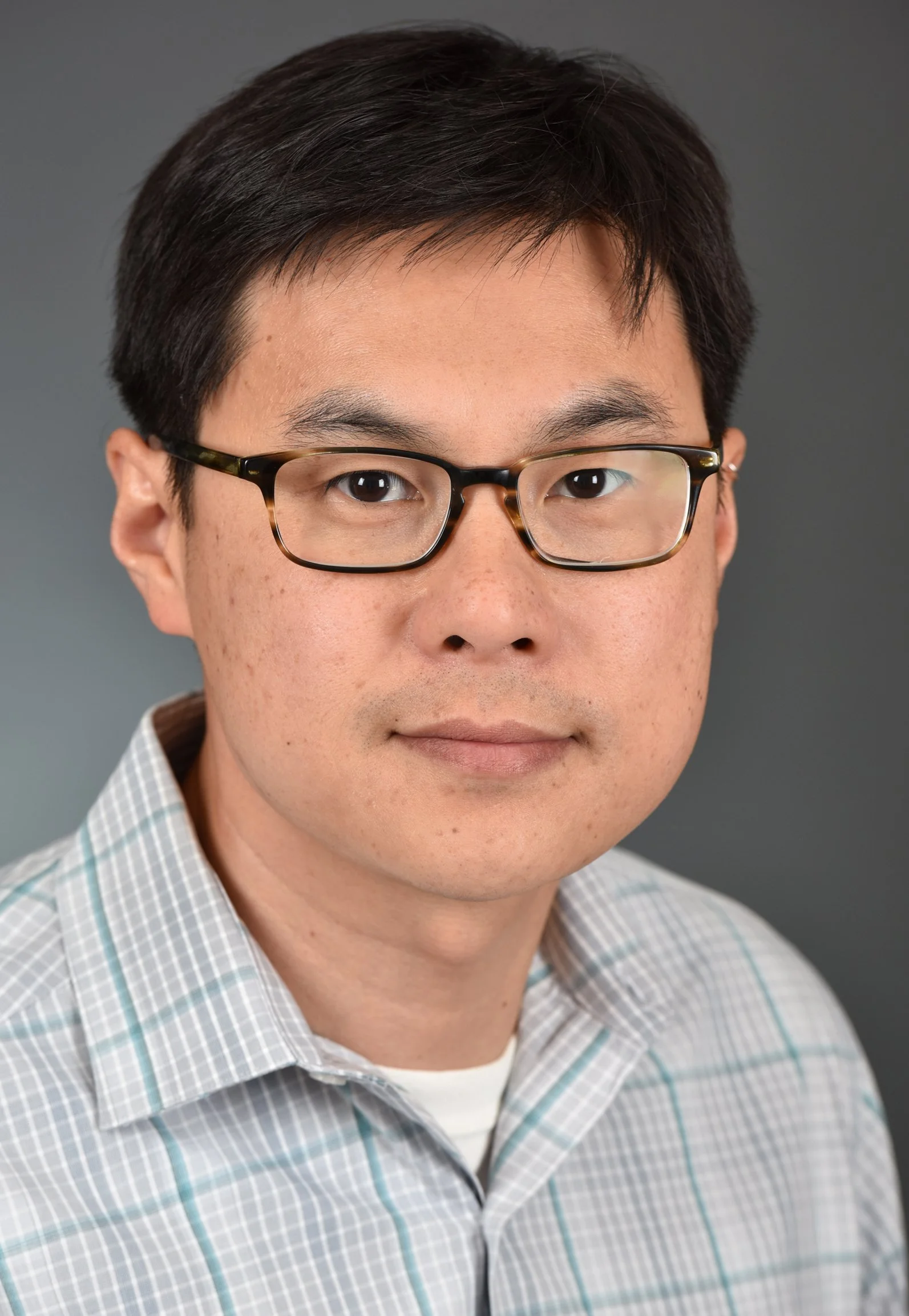 A person with short dark hair and rectangular glasses looks directly at the camera against a plain gray background, wearing a light gray plaid button-up shirt.