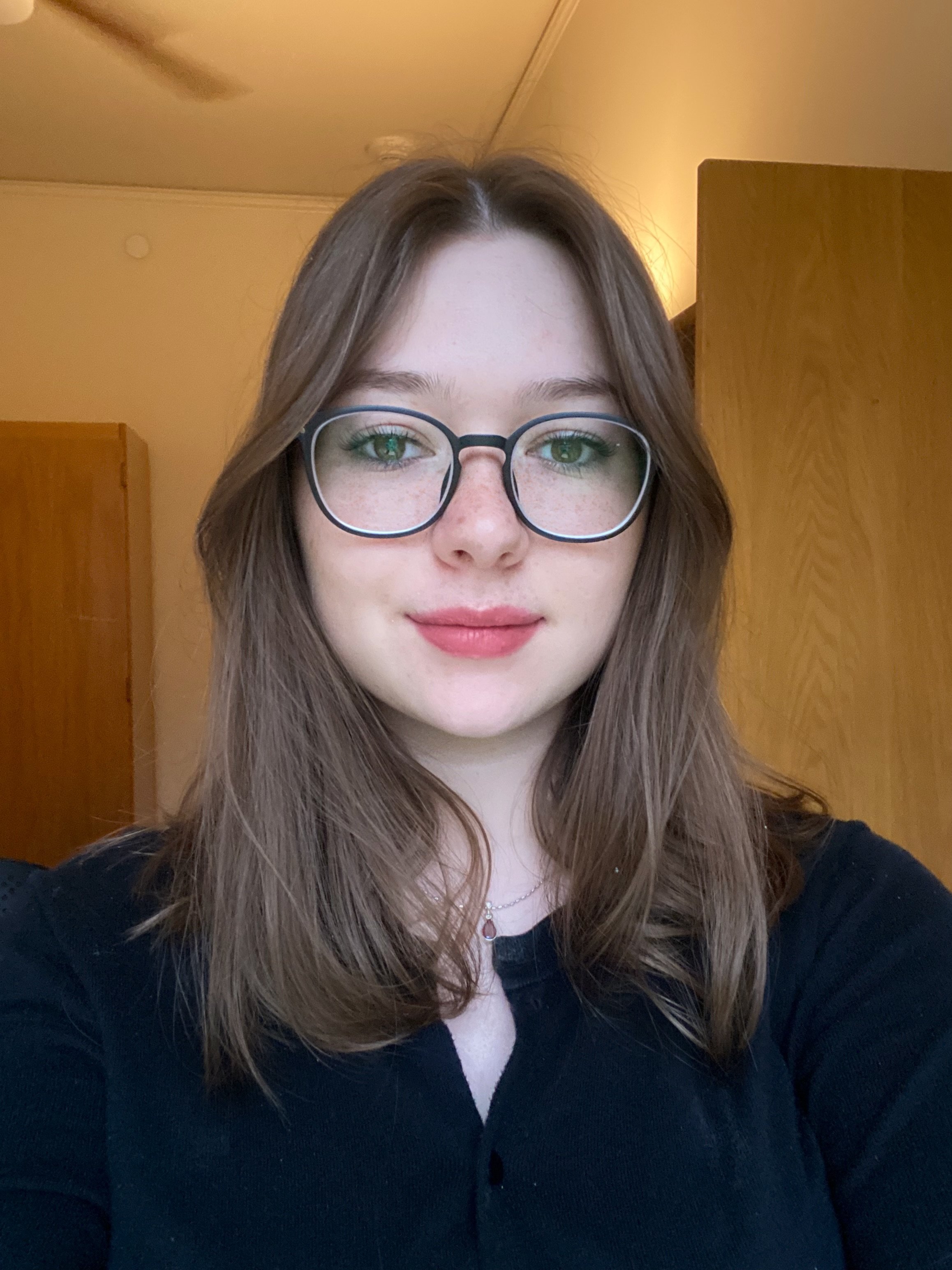 A person with straight, shoulder‑length brown hair and round glasses looks directly at the camera, smiling softly. They wear a black top and a delicate necklace. The background shows a softly lit indoor room with light-colored walls, a wooden door, a