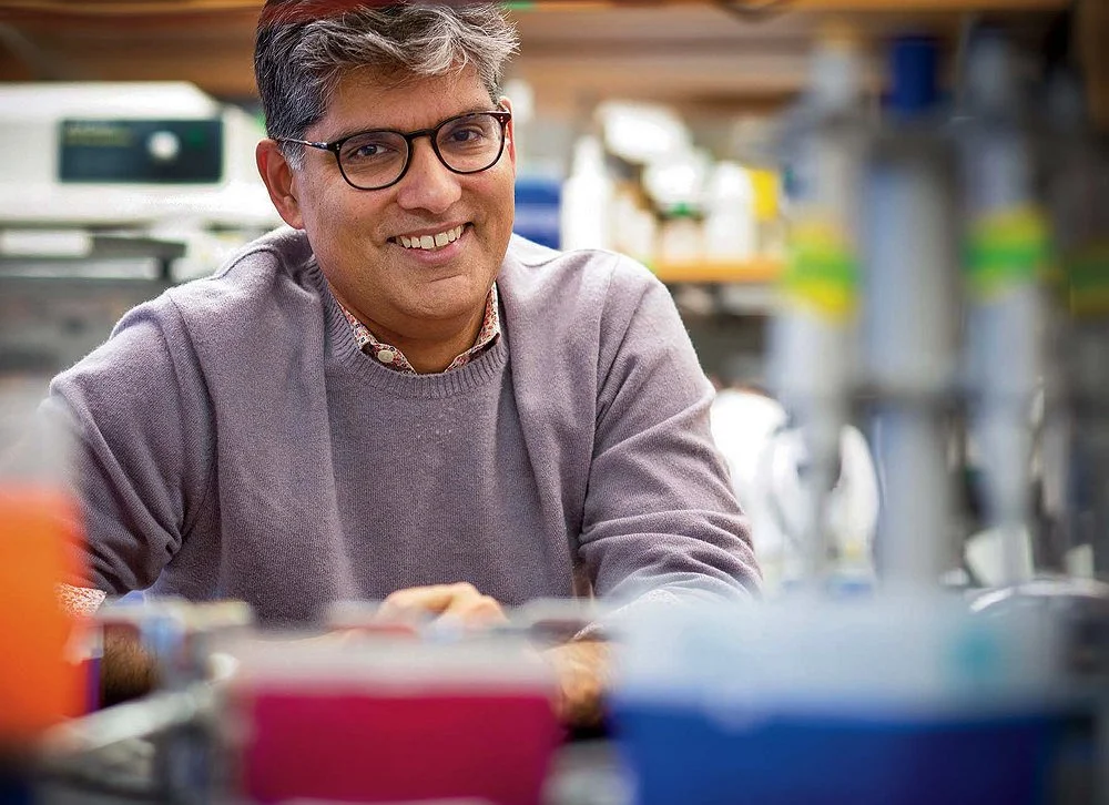 Person with short gray‑streaked hair and glasses smiling in a laboratory, wearing a light purple sweater with blurred lab equipment in the foreground.