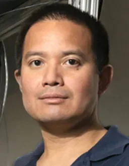 Person with short dark hair looking at the camera with a neutral expression, wearing a dark collared shirt, with blurred equipment and cables in the background.