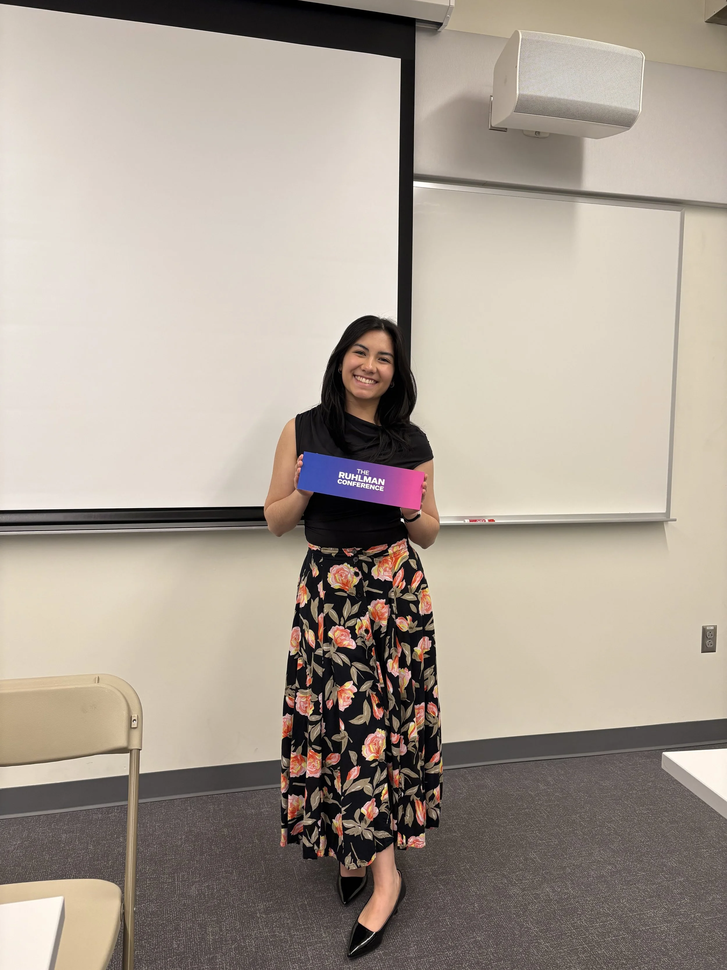 A woman with medium-length hair, wearing a sleeveless black top and long floral skirt, smiling and holding a sign in a classroom with a projector screen behind her.