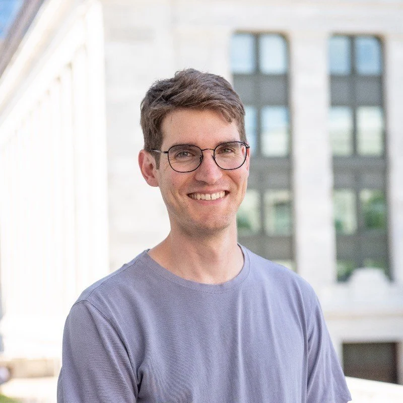 Person with short brown hair and round glasses smiling outdoors, wearing a light gray T‑shirt with a large stone building in the background.
