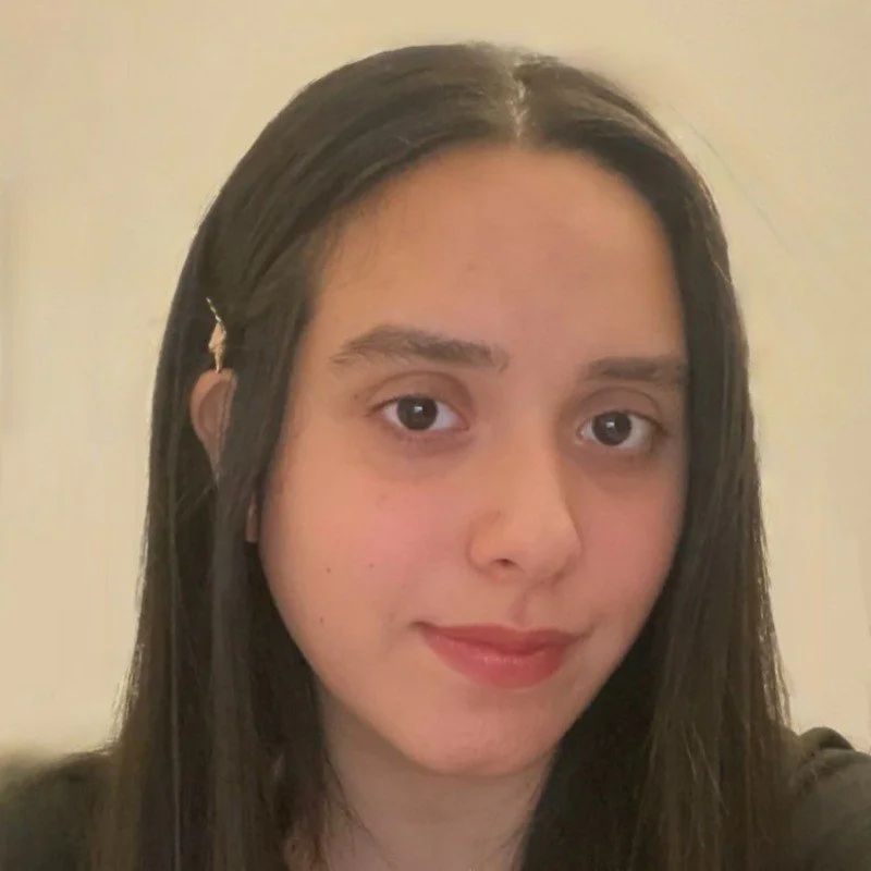 Close‑up of a young woman with straight dark hair parted in the middle, clipped back on one side. She looks at the camera with a neutral expression and wears a soft pink lip color against a plain light background.