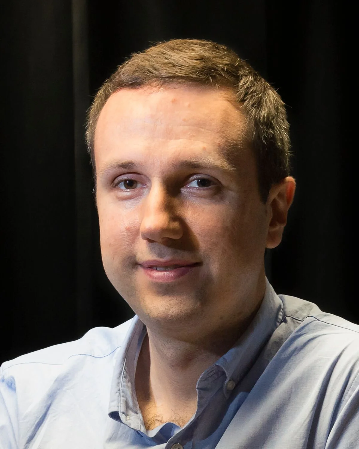 Person with short light brown hair and light skin smiling softly, wearing a light blue button‑up shirt, posed against a dark background.