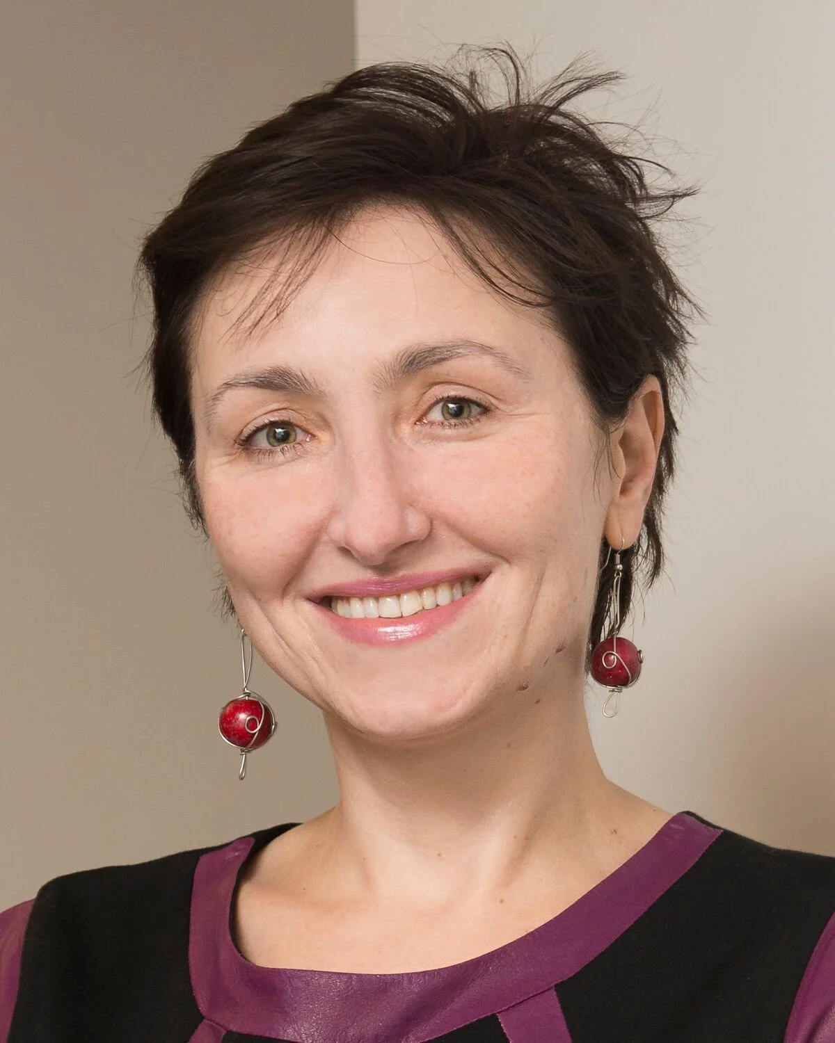 A person with short dark hair and dangling red earrings smiles warmly at the camera, wearing a black top with purple trim, standing in front of a neutral indoor background.