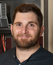 Person with short brown hair and beard smiling slightly at the camera, wearing a dark patterned shirt, with electronic equipment and cables blurred in the background.