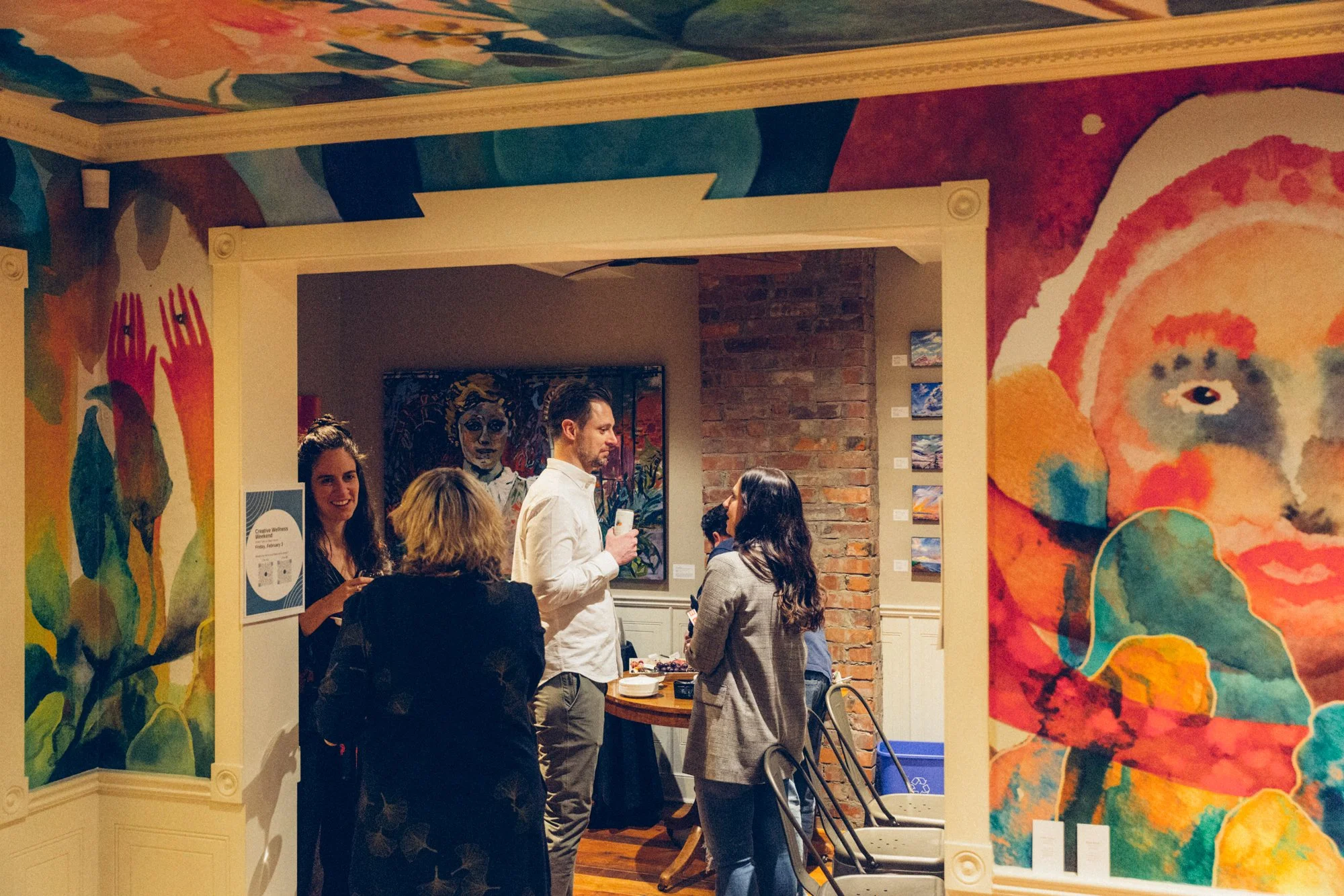 Group of people standing and talking in an art gallery, with colorful paintings and murals on the walls.
