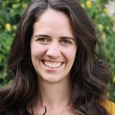 A young woman with long brown hair smiling outdoors with yellow flowers in the background.