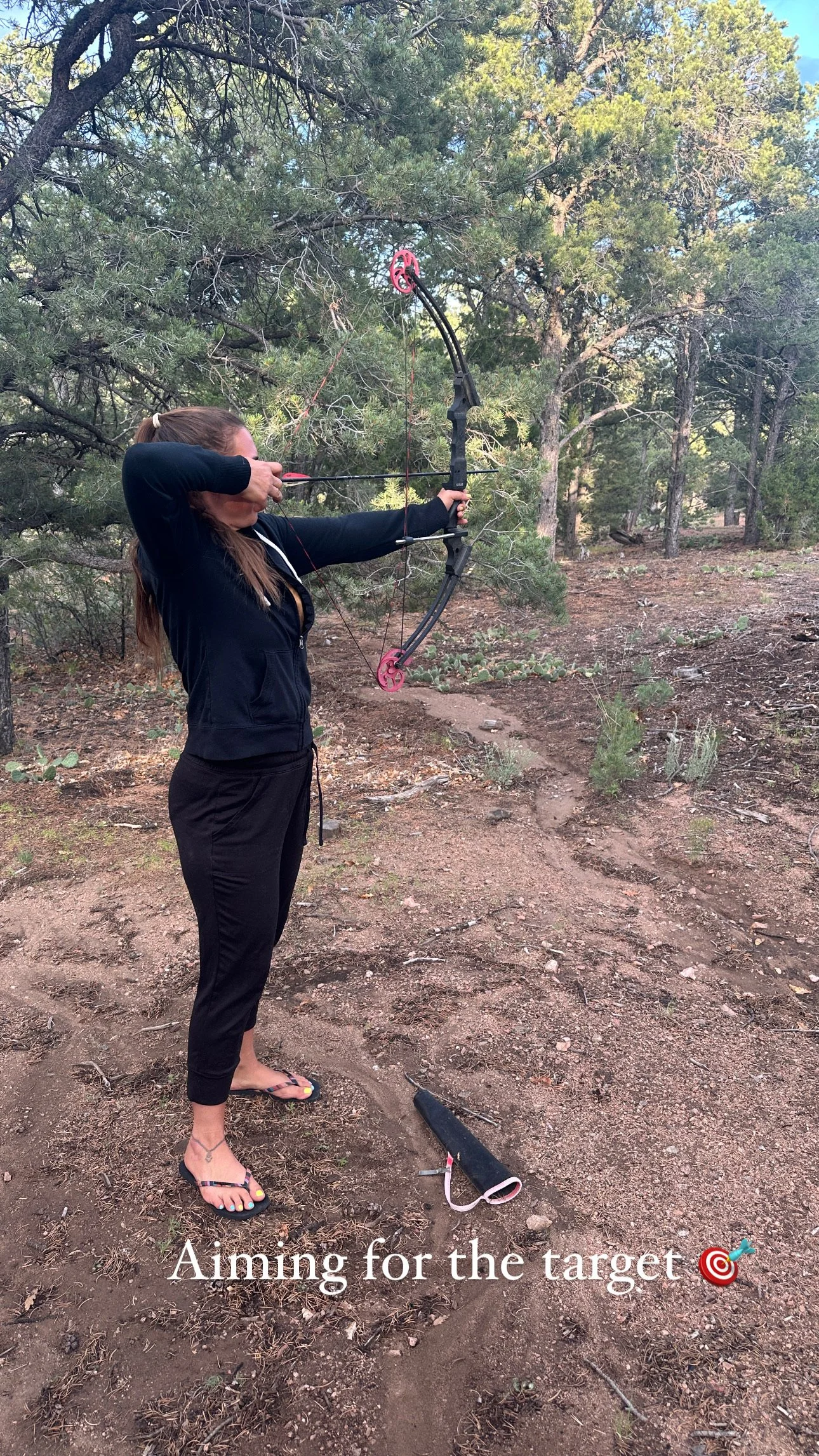 Woman aiming a bow and arrow at a target in a wooded outdoor area.