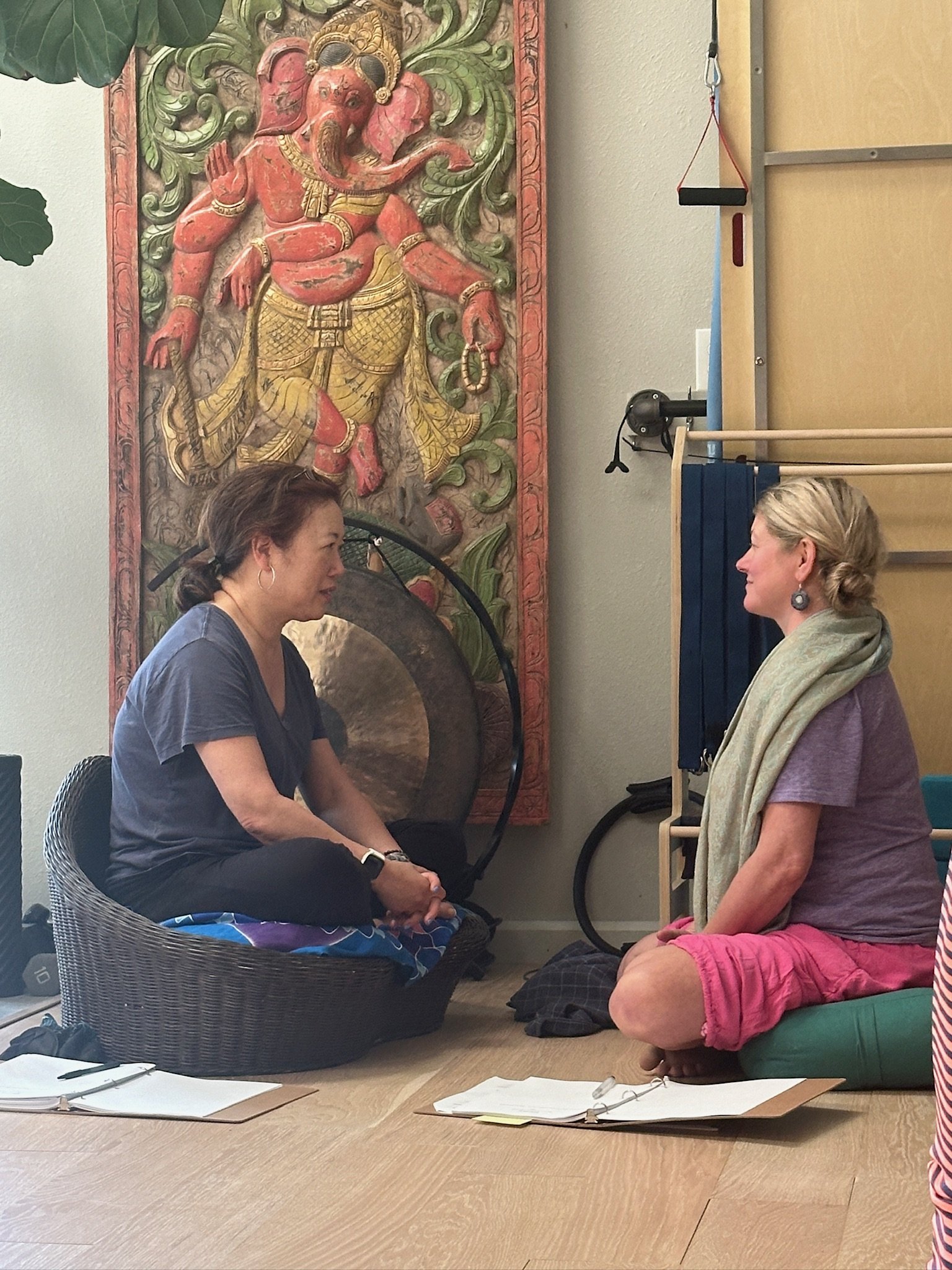 Two women sitting on the floor facing each other with notebooks and pens on a table nearby. There is a large colorful Buddha facing the women on the wall behind them. One woman has dark hair and is wearing a blue shirt, the other has blonde hair and 