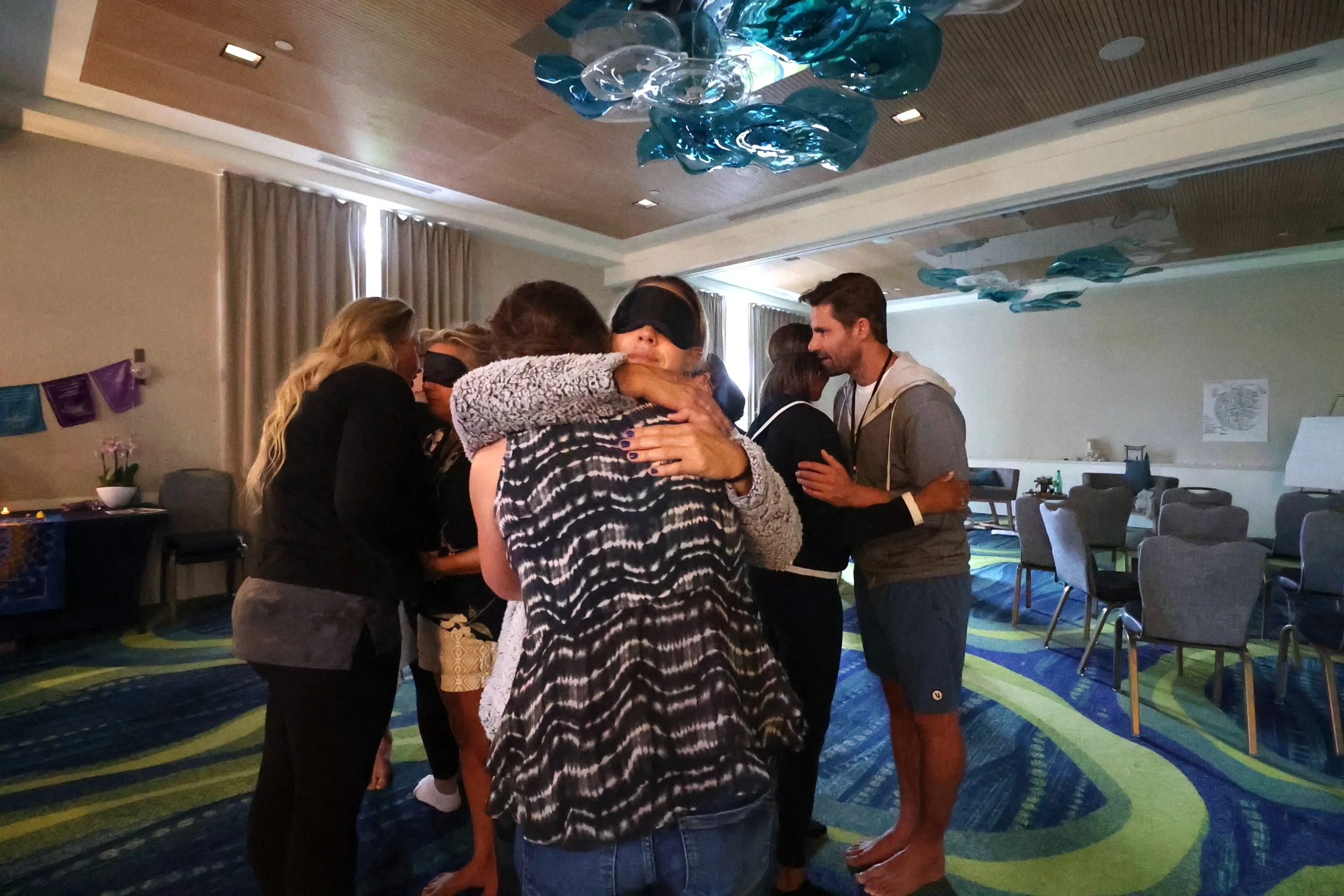 Group of people standing in circle, hugging each other with their eyes closed, in a decorated indoor room.