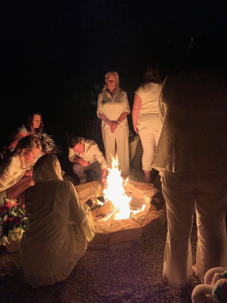 Group of people gathered around a fire pit outdoors at night, some kneeling, some standing, some sitting, with dark sky in background.