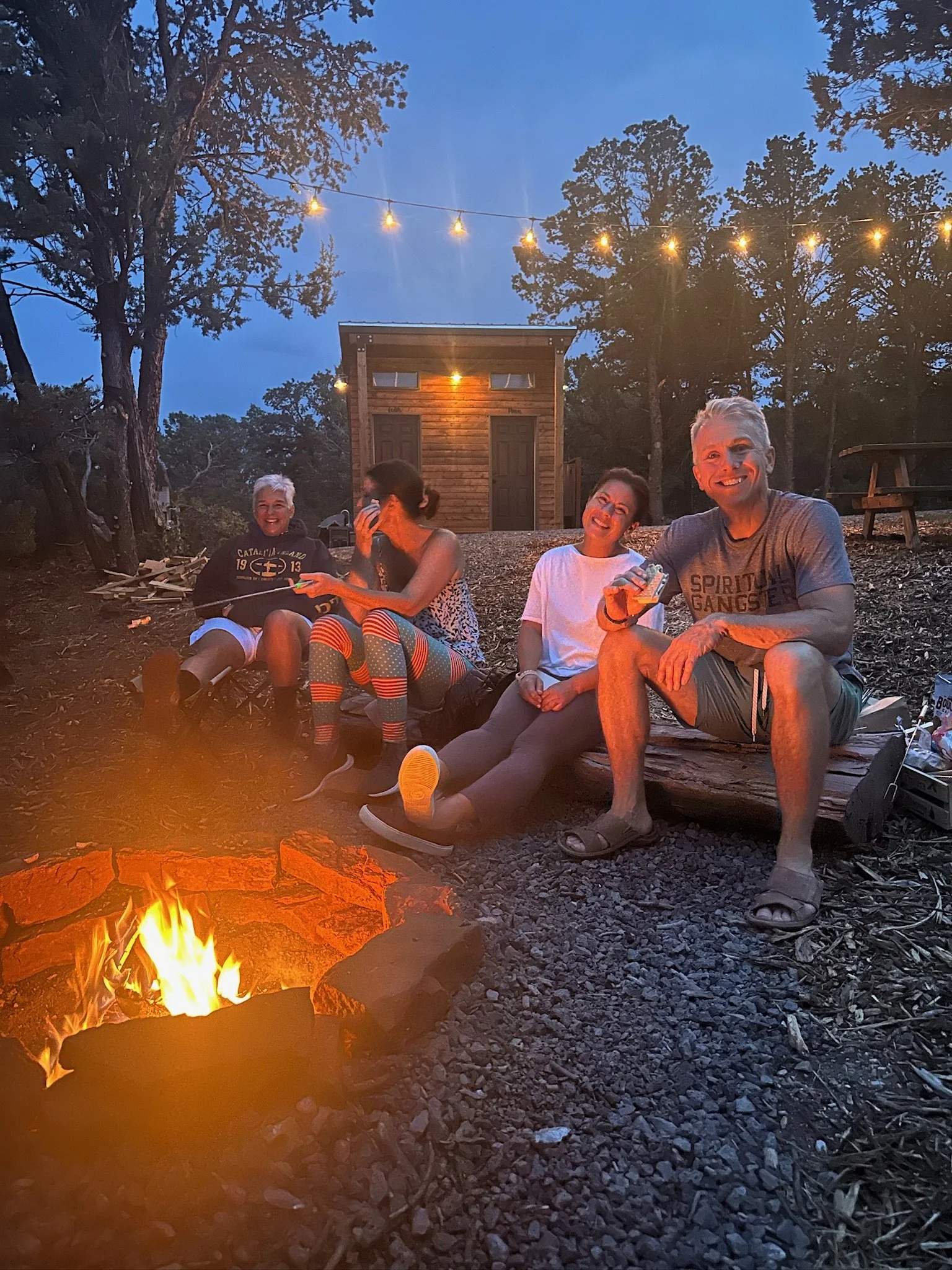 Four people sitting around a campfire at dusk in a wooded outdoor area with string lights overhead. They are smiling and enjoying each other's company.