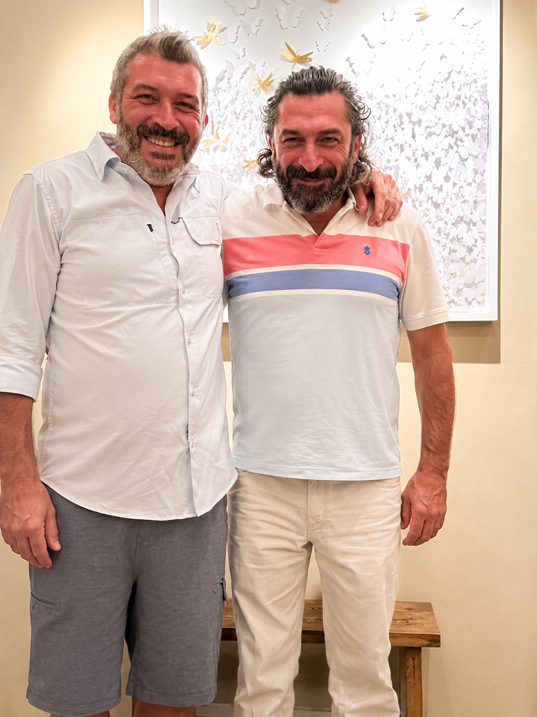 Two smiling men standing close together, with one man's arm around the other's shoulder, in front of a decorative wall with butterfly patterns.